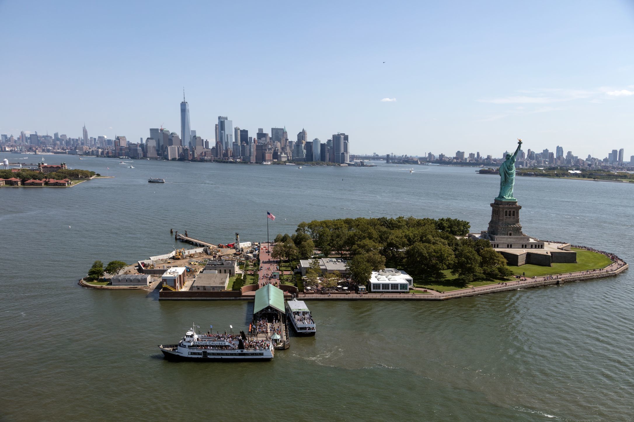Aerial view of Liberty Island with buildings, trees, the Statue of Liberty, and the city over the water in the background.