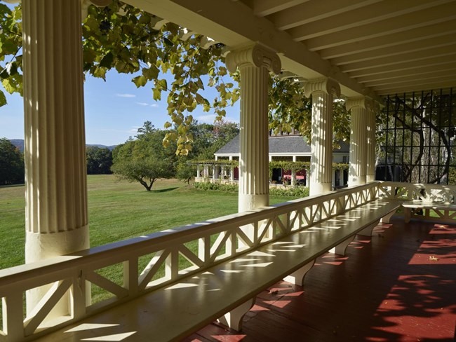 View of the Little Studio from the porch at Aspet Vines frame a view through columns of a porch, towards a grassy lawn, an apple tree, and a studio surrounded by a pergola.