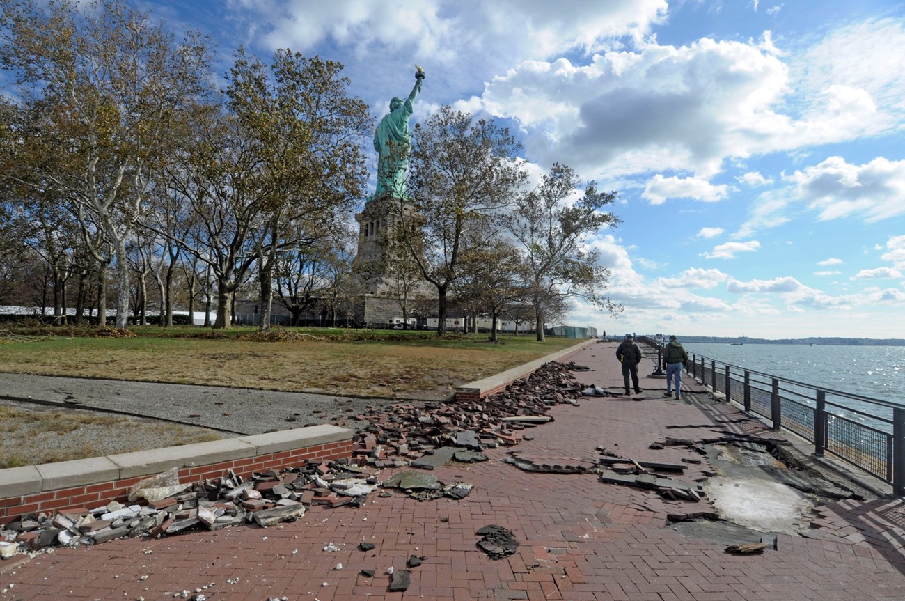 Broken bricks from storm damage are scattered along a walkway with the Statue of Liberty in the background