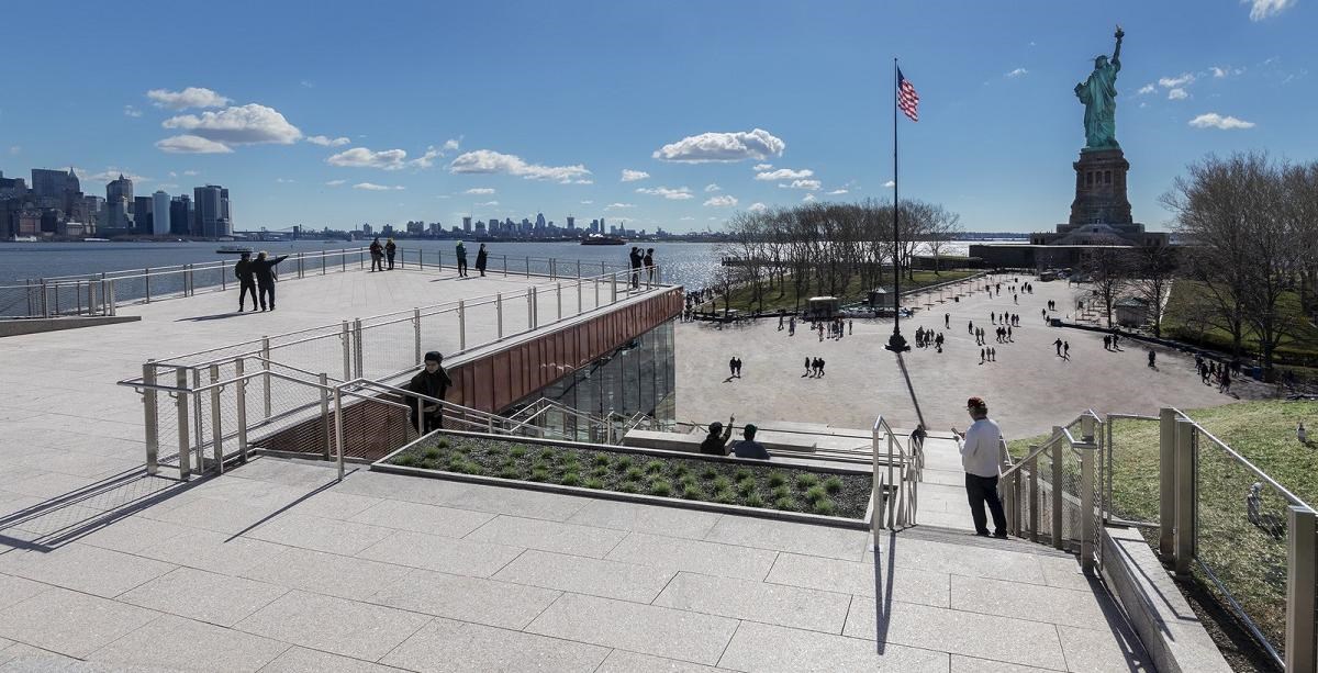 Statue of Liberty Museum view A viewing platform faces the water and city skyline to the left and Statue of Liberty beyond a round plaza with a flag in the center.