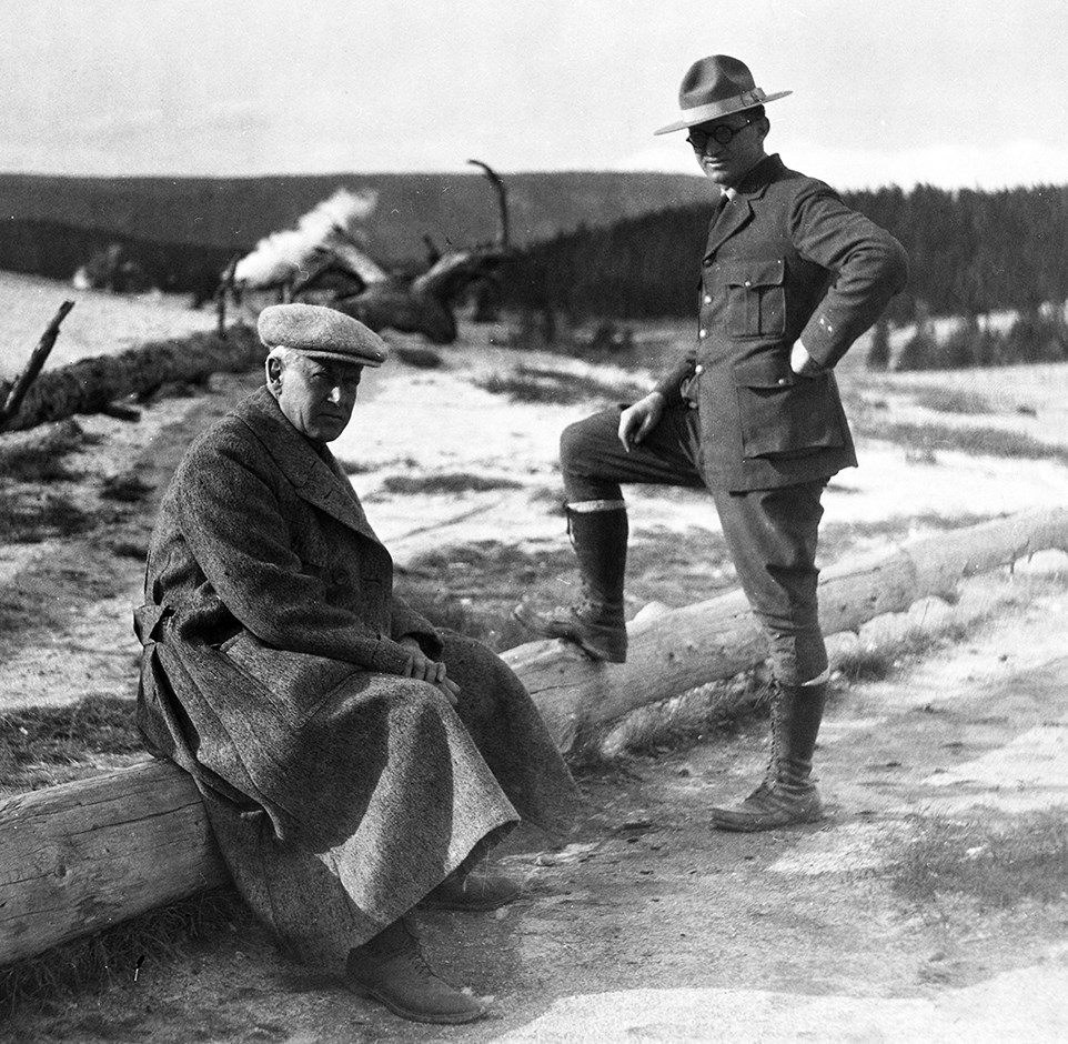 A man in NPS uniform stands with one foot on a fallen log, beside a seated man in a long coat and cap.