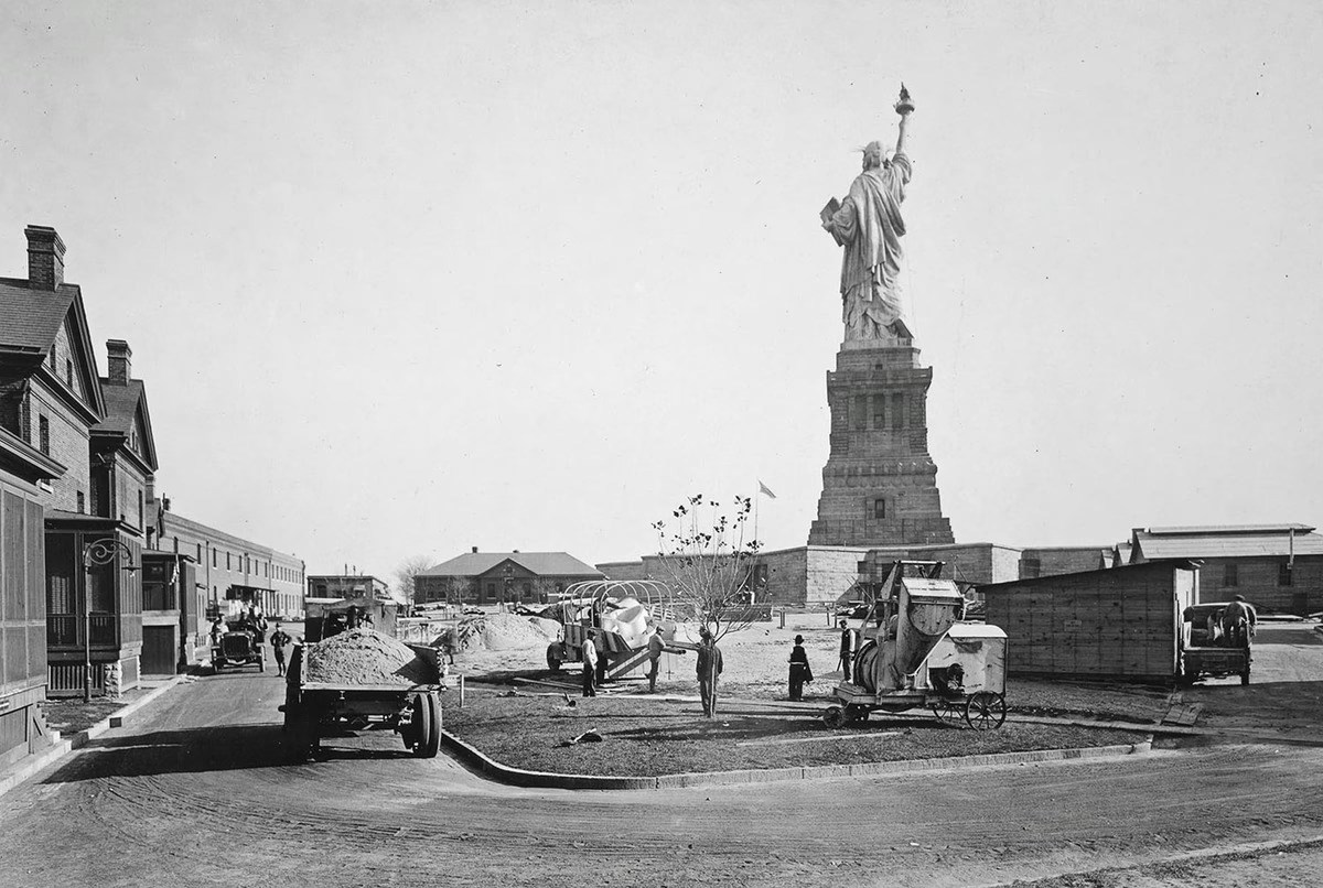 Workers use vehicles to move dirt in front of buildings at Fort Wood, with the Statue of Liberty in the background.