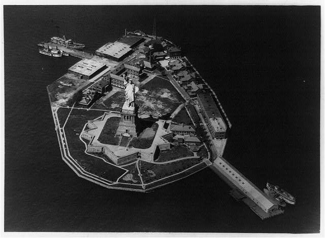 Historic aerial photo of Liberty Island showing the arrangement of buildings, landscape, Statue of Liberty, and surrounding water.