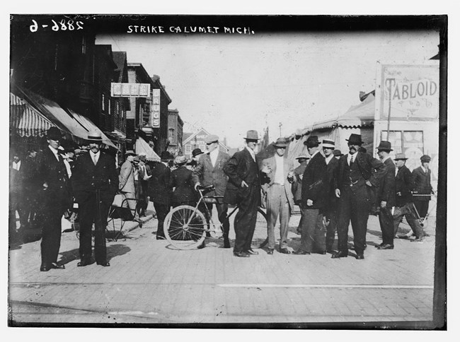 Men in suits and hats gather in a city street that is lined by low buildings, between 1910 and 1915.