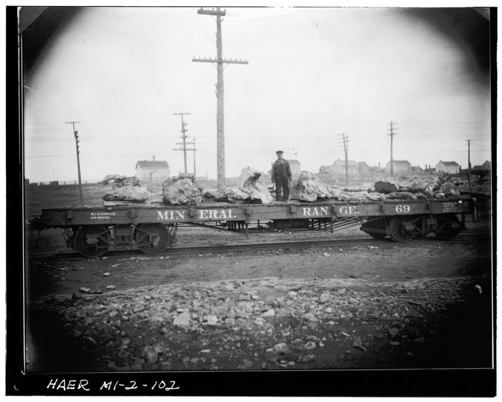 A man stands on a railroad flatcar, alongside large masses of rock or copper. Telegraph poles and scattered houses stand in the background.