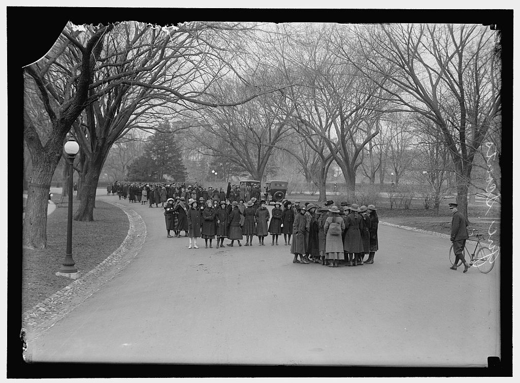 Groups of girls in long coats gather on a curving road, flanked by leafless trees, streetlights, and stone guttering