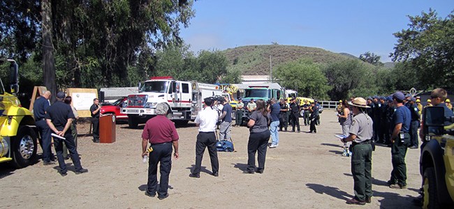 lt= A group of press members and NPS employees stand in an open parking lot during a press conference.