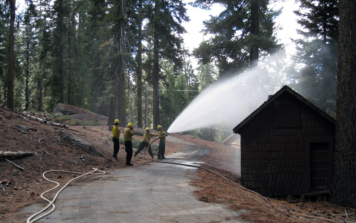 lt= A firefighter with a hose sprays a building's roof, while three other firefighters stand by.