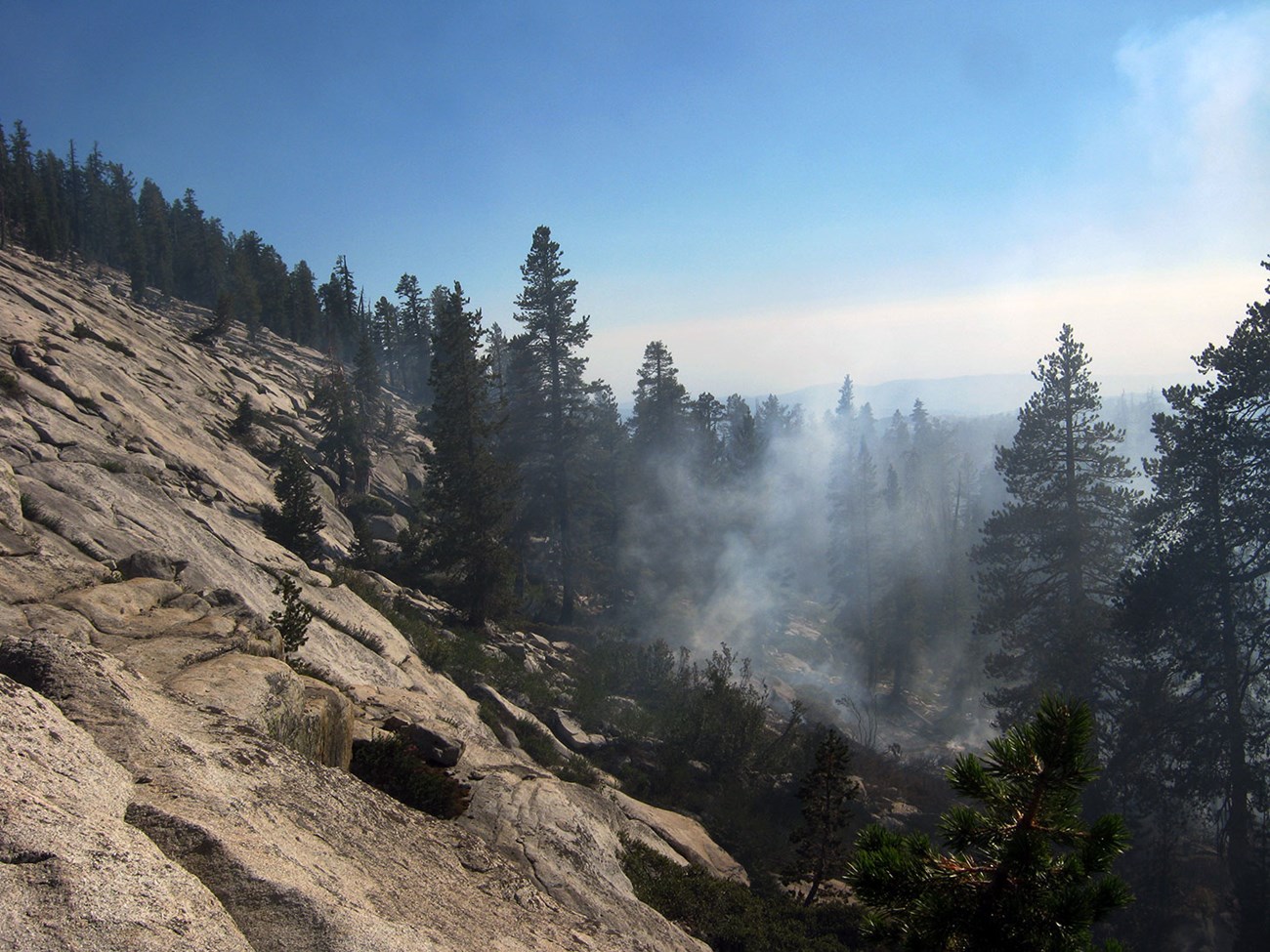 Light smoke rises from a forest near a bare rock face.