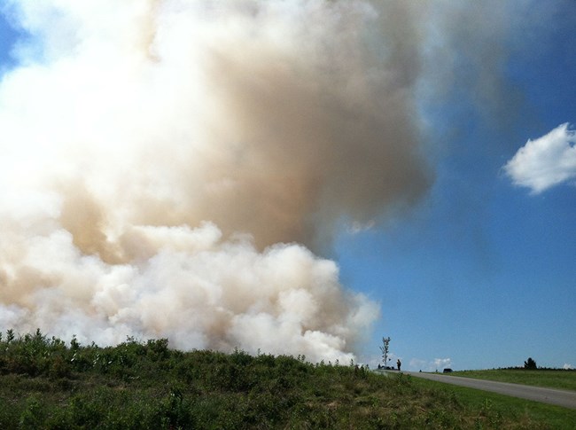 A large plume of smoke rises from behind a small hill adjacent to a road.