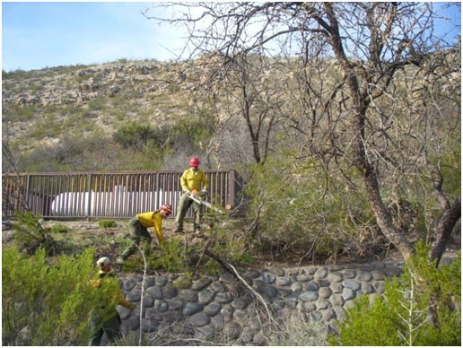 lt= Three firefighters work near a stone wall and propane tank in a desert environment.