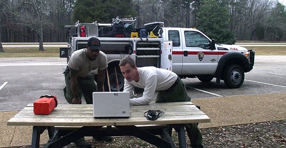 Two firefighters use a laptop on a picnic table while a wildland fire engine is parked behind them. Two firefighters use a laptop on a picnic table while a wildland fire engine is parked behind them.