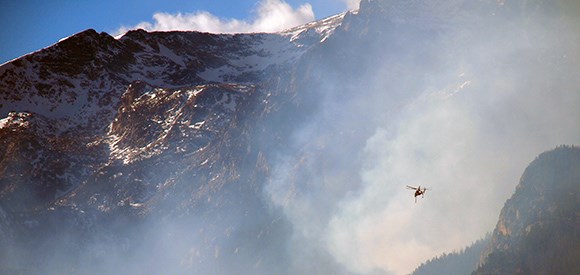 A Type 1 helicopter flies out of a smoky drainage with a craggy mountain in the background.
