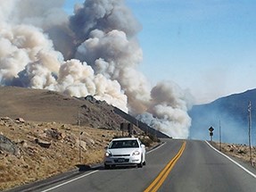 Large plume of smoke behind hill as car drives on road toward camera.