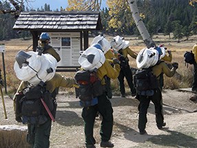 Firefighters start down a hiking trail with coils of hose on their backs.