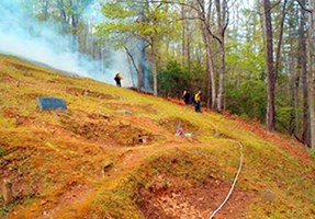 Smoke and firefighters at edge of trees on slope, with gravestone in grassy area in foreground.