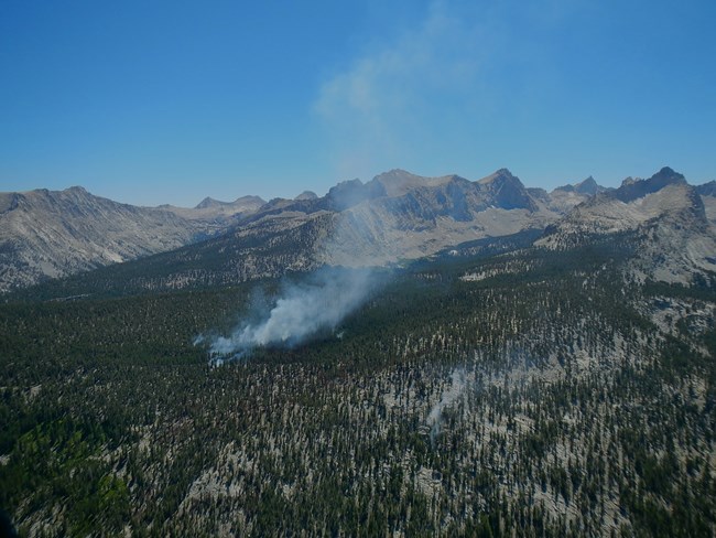 A wilderness fire is in a mixed conifer forest with white smoke coming up from it in the middle of the image. In the distance, barren high mountain peaks are seen.