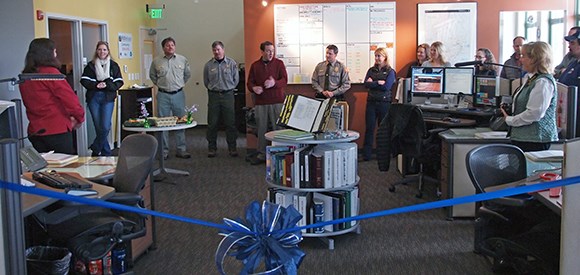 A group is gathered in a room with a ribbon waiting to be cut in front.