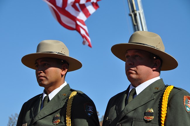 National Park Service Fire and Aviation Honor Guard Two men in dress NPS uniform stand beneath a United States flag.