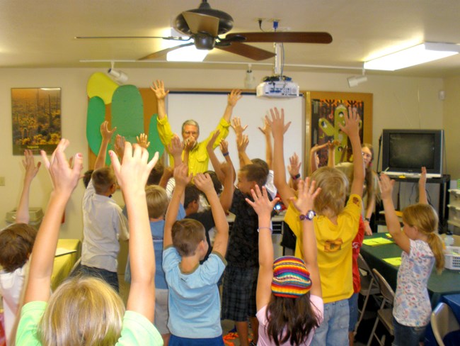 A firefighter talks with students in a classroom. All their arms are raised in a stretch.