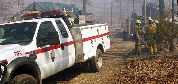 Two firefighters in Nomex stand near an NPS engine on a wooded property.