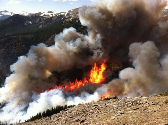 Flames and dark smoke on a dirt and rock slope with snow-capped mountains in the distance.