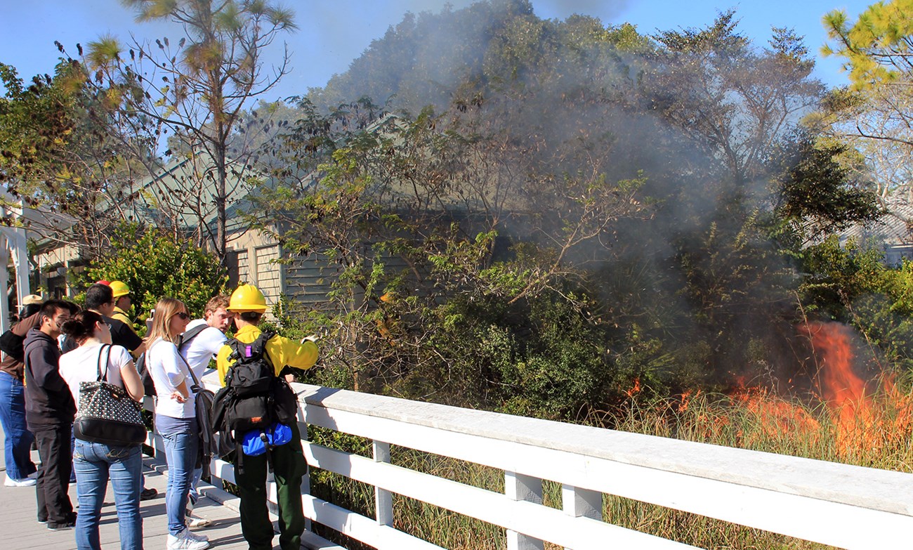 A firefighter talks to visitors along a boardwalk while fire burns on the other side of a fence.