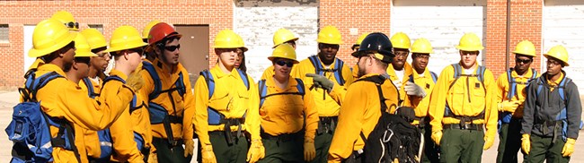 Handcrew in training A group of young men and women in firefighting gear listen to a man speaking to them.
