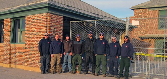 A group of men stands in front of red brick buildings with green trim.