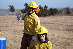 Two crew members, one with a chainsaw, the other kneeling, smile at the camera.