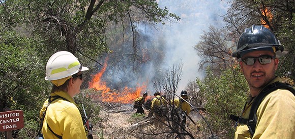 A man and woman dressed in Nomex flame-resistant clothing stand in the foreground near a sign for Visitor Center Trail, while several firefighters work near a fire in the background.<