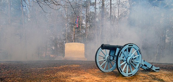 lt= A monument stone is obscured by smoke and surrounded by burned vegetation, while a cannon stands watch in the foreground.