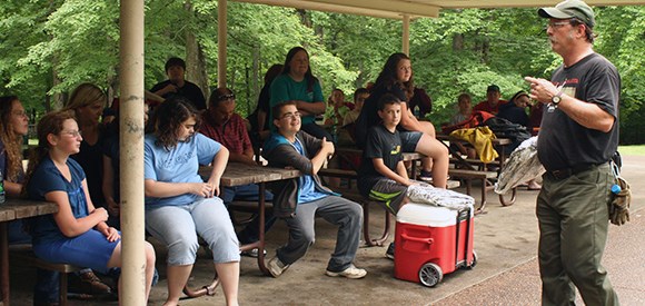 Fire Education Kids sit on picnic tables under a shelter listening to a firefighter.
