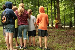 Hot Shots campers stand and watch a chainsaw demonstration taking place near a grove of trees.