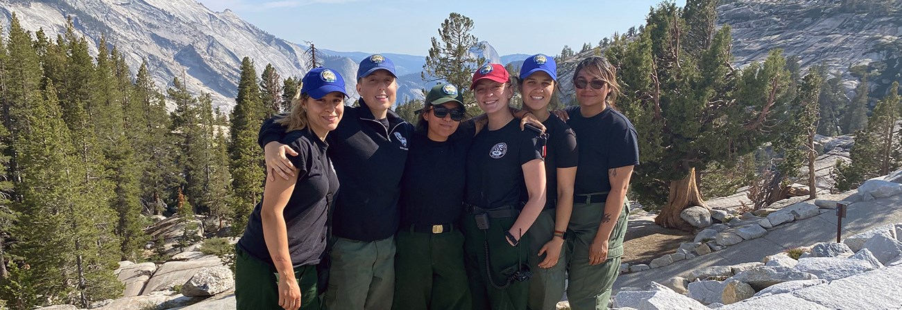 A group of six women in blue shirts and green pants stand on a scenic overlook with Half Dome in the background.