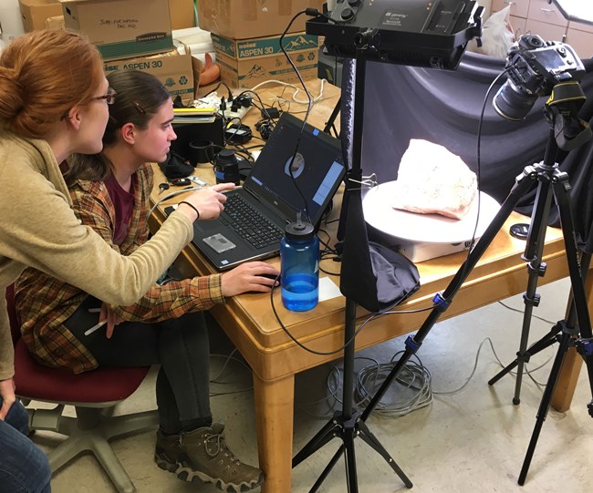 two people work at a computer with camera and lights set on rock sample