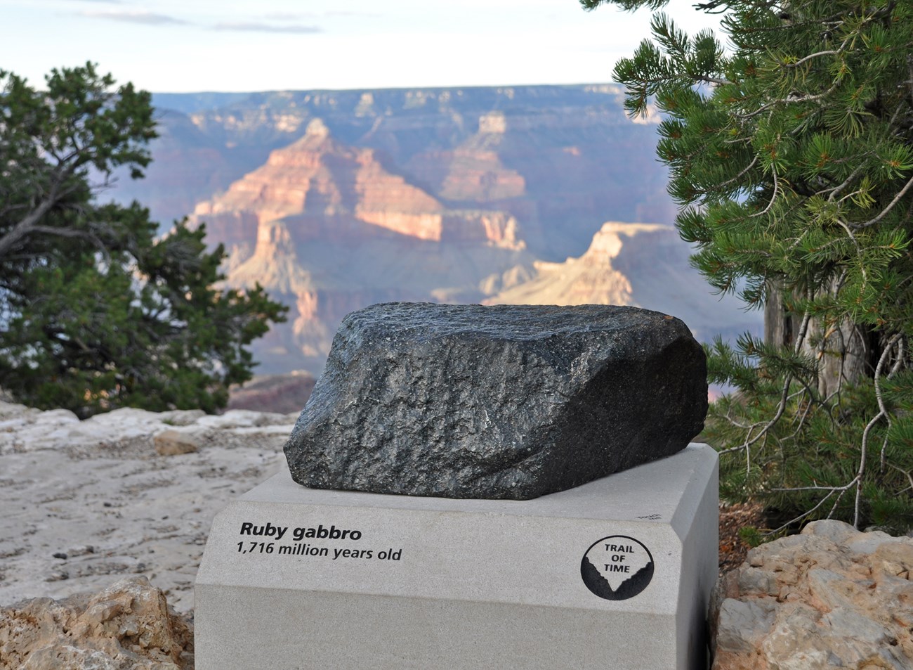 A dark color boulder displayed outdoors with the Grand Canyon in the distance.