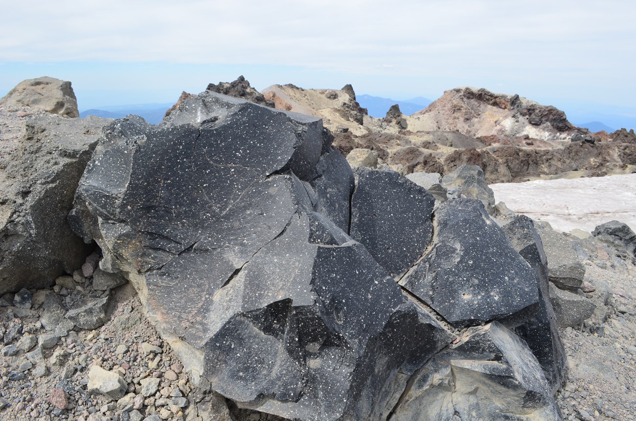 A shiny black dacite boulder near the summit of Lassen Peak, with patches of snow in the background.