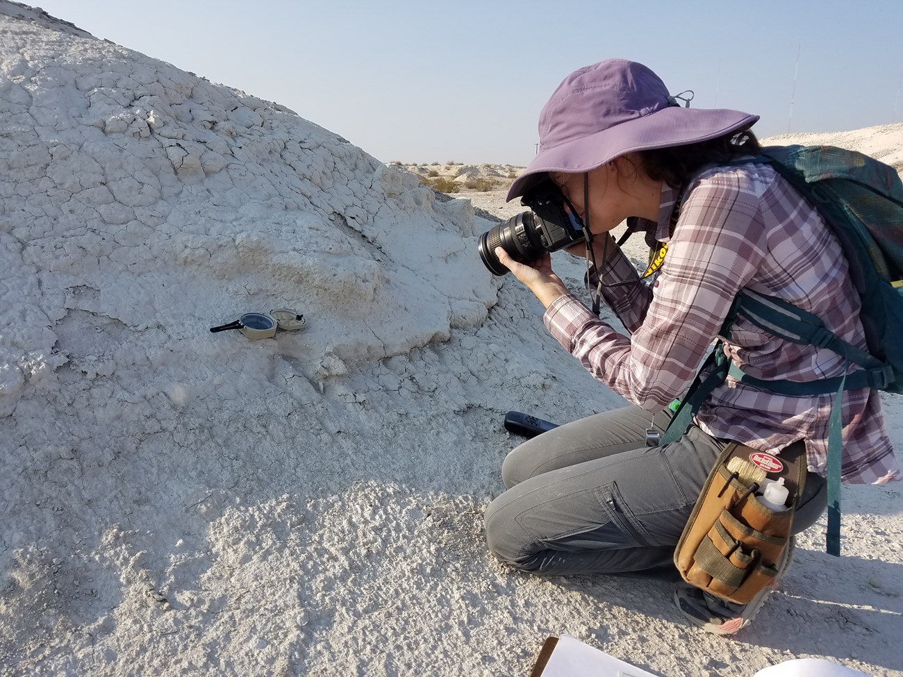 A young woman in a purple hat and sunshirt kneels next to a rock. She looks through her digital camera to photograph a fossil.