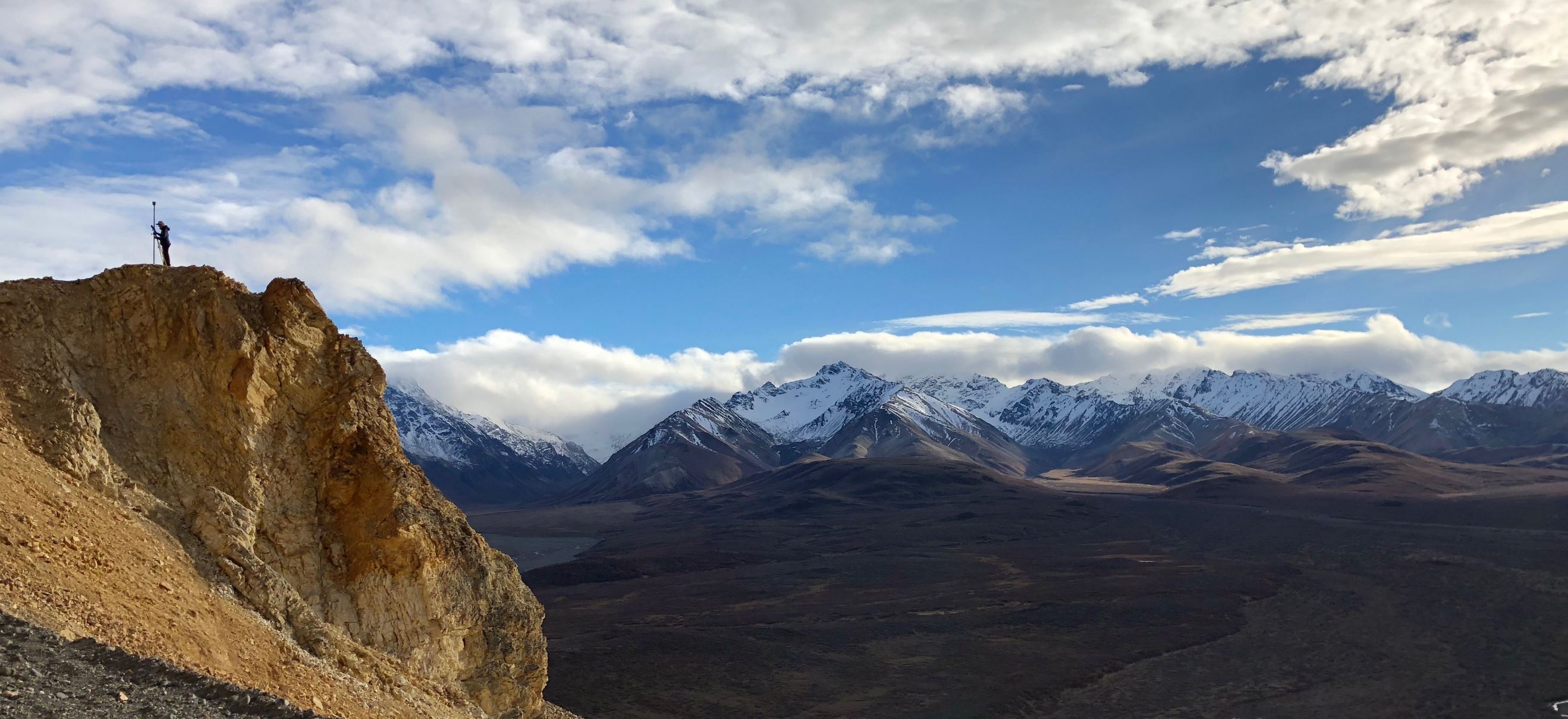 View over large valley in Denali National Park, with snow capped mountains in the background, and a person performing GPS surveying on a cliff edge in the foreground.