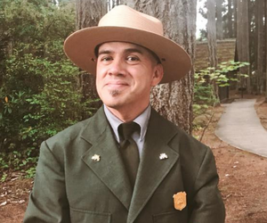 Learn more about Cam Juarez. picture of NPS park ranger posing in uniform with nature and trail in background
