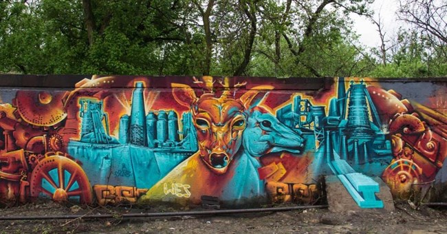 Carrie Deer Head Mural An orange deer head and a blue deer head sit in front of blue iron works. A mural.