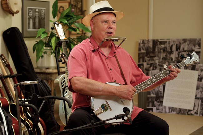 An older white man in a pink shirt and jeans plays a banjo and sings into microphones