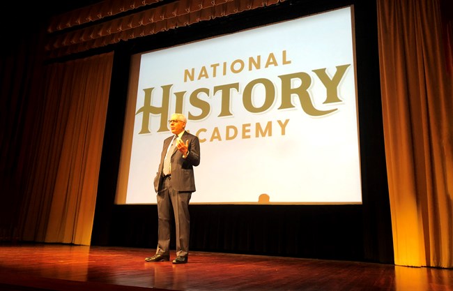 A man stands on a stage in front of a projection screen that says "NHA Academy"