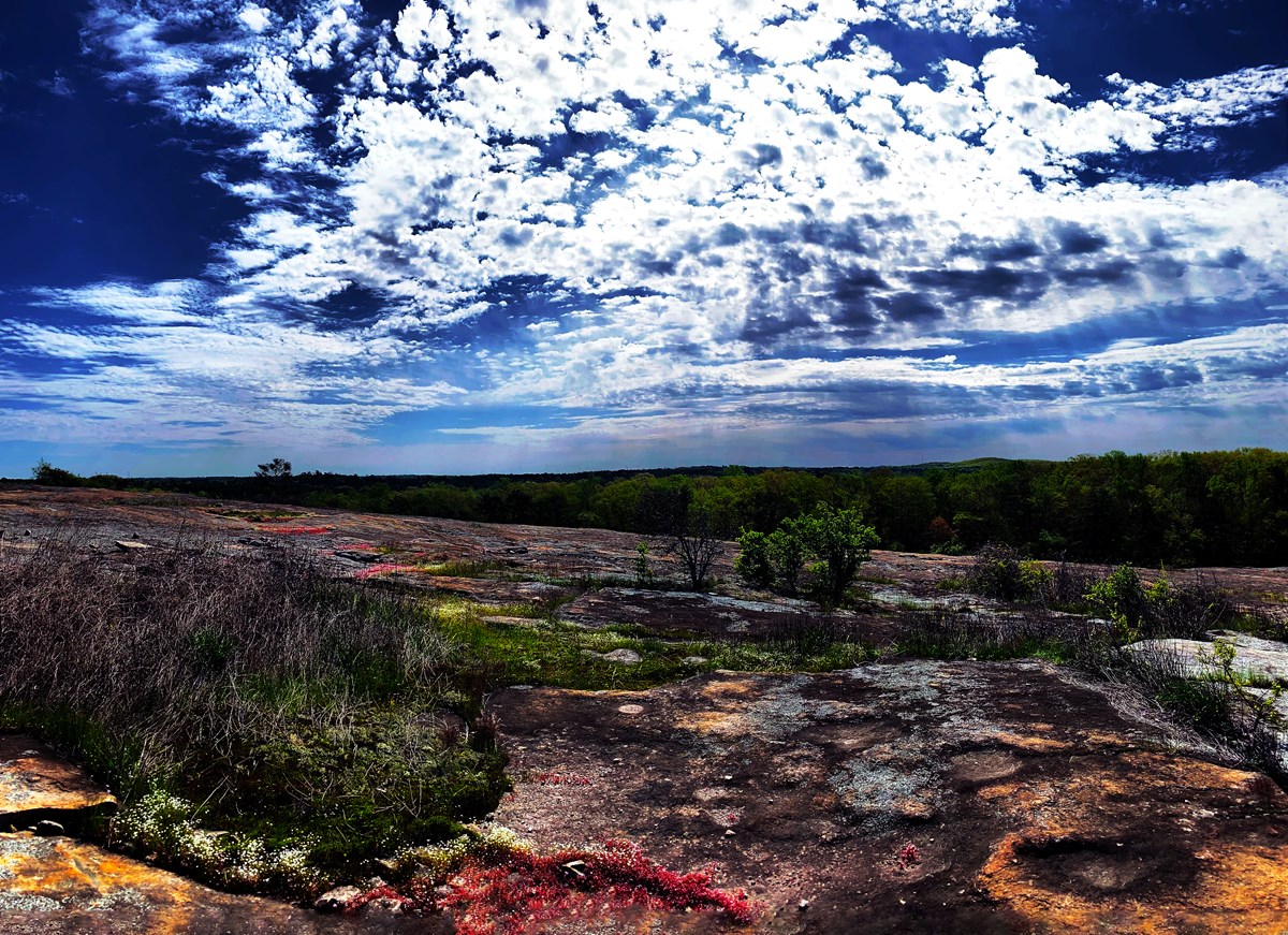 View of the granite top of Arabia Mountain, with red and orange growths, under a blue sky and dramatic clouds.