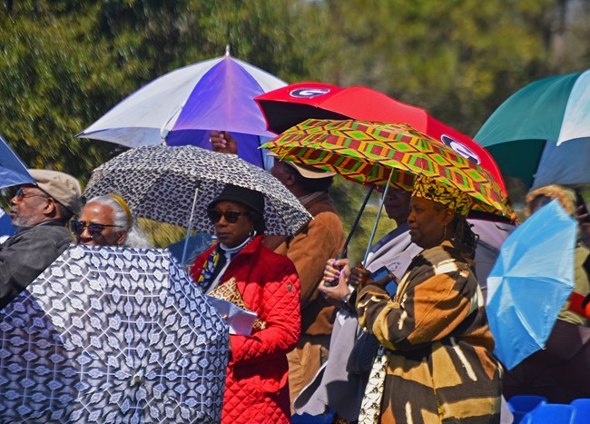 Attendees carry multicolored, multi-patterned umbrellas.