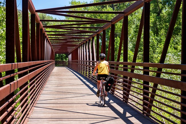 A person bikes across a suspension bridge. Metal beams of the bridge cast diamond patterned shadows.