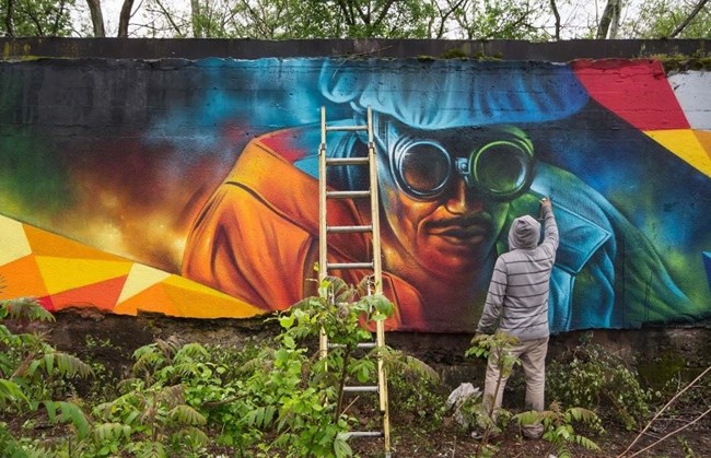 A mural painting, in progress A mural of a blue iron worker being painted by a man in grey next to a ladder