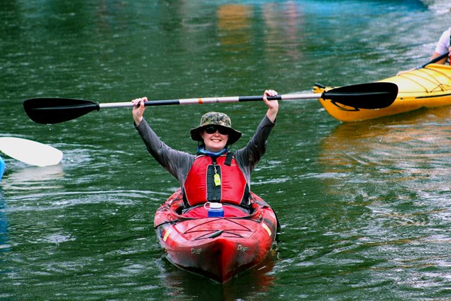 A smiling woman in a red kayak raises her paddle above her head victoriously.