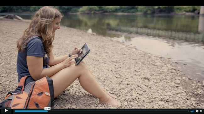 A woman sits on a pebbly river bank looking at an ipad.